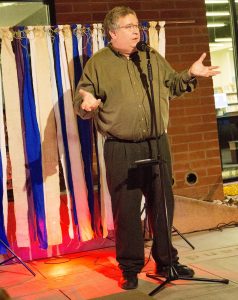 Sean Buvala stands in front of a cloth strips stage set. he is wearing a green shirt and is in front of a microphone. It is important when you want to have a conversation that you know your stories and storytelling